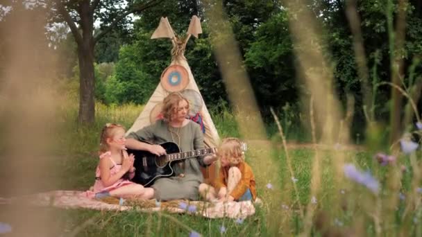 Mom playing the guitar to her little daughters next to wigwwam outdoor in summer — Stockvideo