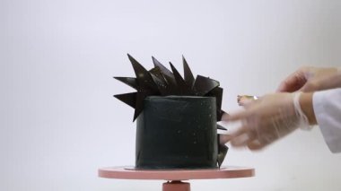 Profesyonel pasta şefi, acımasız bir siyah pastayı pembe bir tezgahta çikolata dilimleriyle süslüyor. Beyaz arkaplanda fütüristik tarzda tatlı