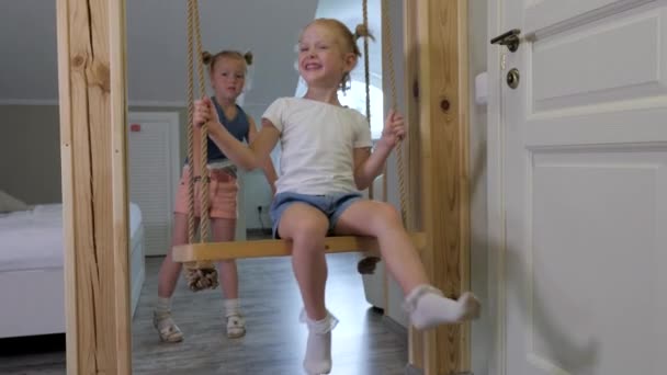 两个可爱的金发碧眼的带着雀斑的姐妹骑在家里的木制秋千上 儿童的家庭娱乐 无忧无虑的童年 — 图库视频影像