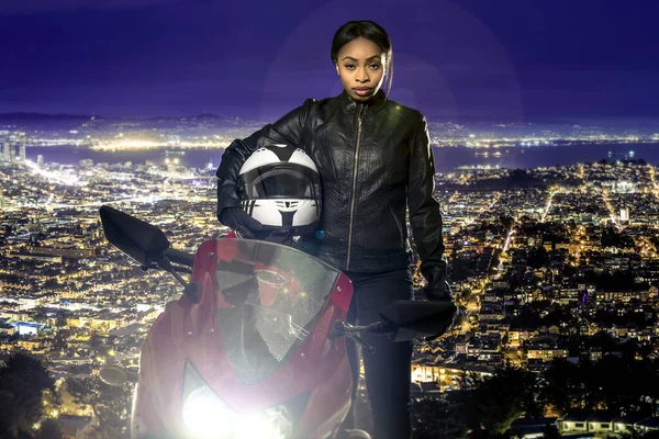 Sporty female motorcyclist or biker wearing a leather jacket and helmet on a road at night with a motorcycle above a scenic view of the San Francisco cityscape