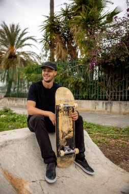 Gülümseyen beyaz bir erkek sokakta elinde kaykayla oturuyor.