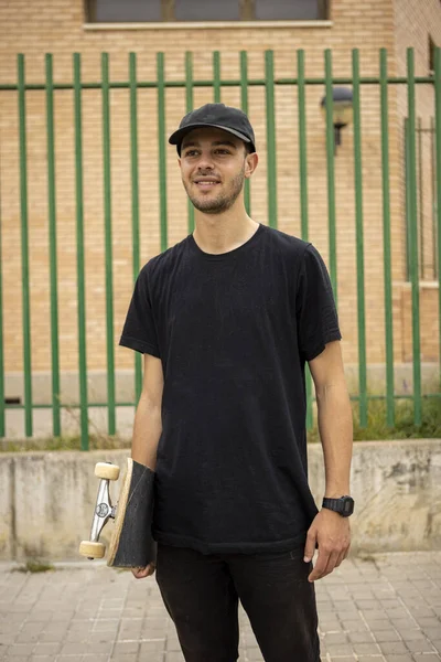 Joven macho con camisa negra y gorra sosteniendo un patín en la mano en la calle — Foto de Stock