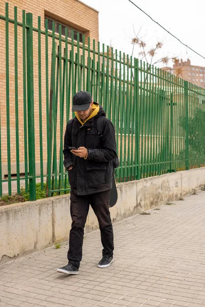 Joven hombre con un abrigo negro y gorra caminando por la calle mirando el teléfono — Foto de Stock