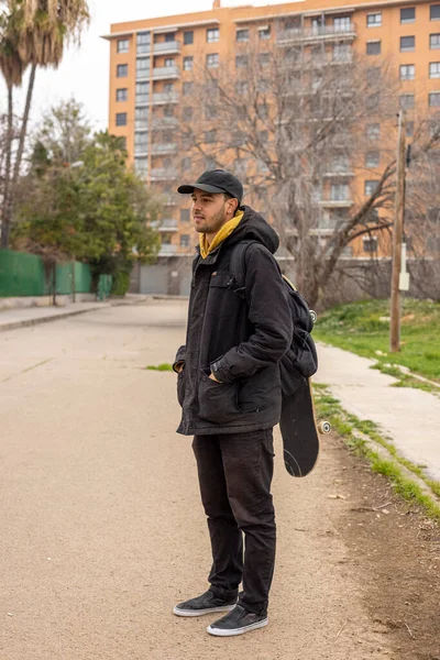 Joven hombre con un abrigo negro y gorra de pie en la calle con un monopatín — Foto de Stock