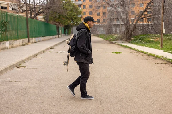 Joven macho con un abrigo negro y gorra caminando por la calle con un monopatín — Foto de Stock