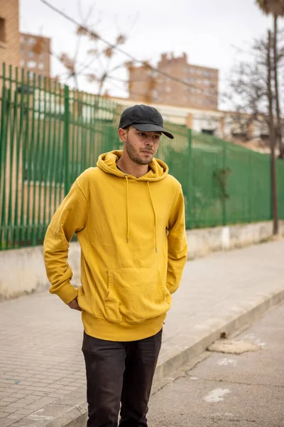 Jeune caucasien mâle dans un sweat à capuche jaune et casquette noire debout dans la rue Images De Stock Libres De Droits