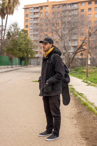 Jeune homme avec un manteau noir et un bonnet debout dans la rue avec une planche à roulettes Photos De Stock Libres De Droits