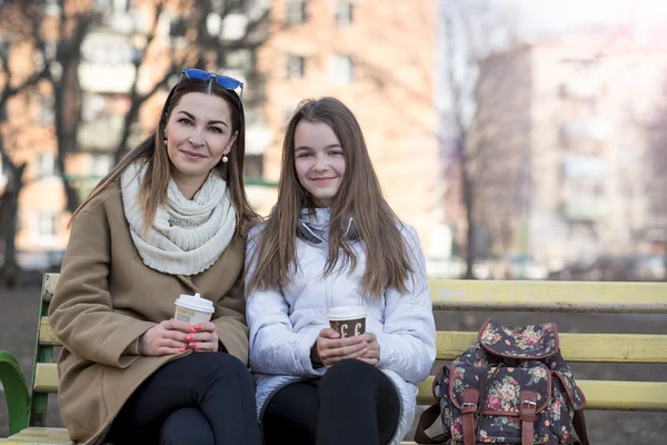 Madre e hija sentadas en el banco del parque de otoño o primavera, se mantienen las manos de las tazas de café —  Fotos de Stock