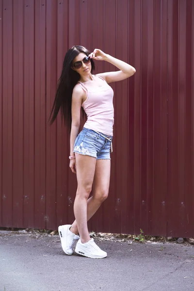 Девушка в очках шортах и розовой блузке стоит перед деревянным забором, стильные модницы . — стоковое фото