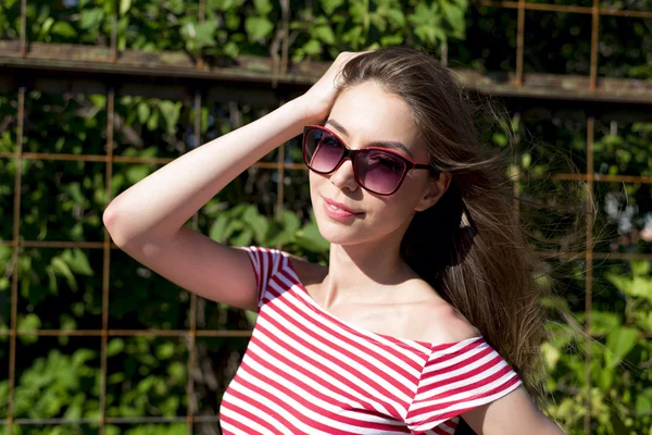 Hermosa chica en una camiseta de color rojo brillante, estilo de vida de moda, posando contra el telón de fondo de la cerca con hojas verdes — Foto de Stock