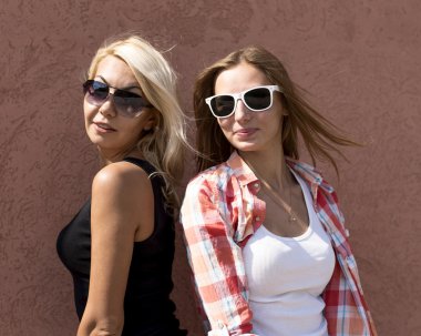 Two girls posing on a brown background wall, glasses, blonde and brunette
