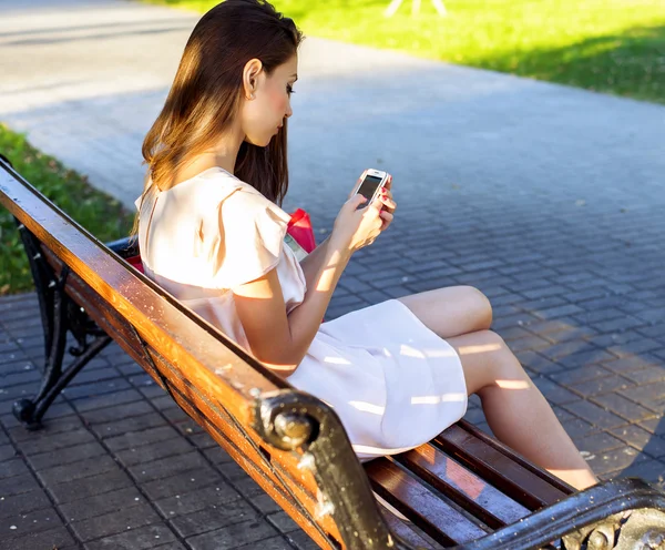 Bella ragazza seduta panchina, abito rosa bruna, stile di vita alla moda con il telefono scrive un messaggio sui social network — Foto Stock