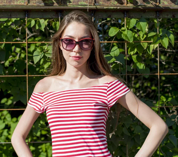 Schöne Mädchen in einem leuchtend roten T-Shirt, Mode-Lifestyle, posiert vor dem Hintergrund des Zauns mit grünen Blättern — Stockfoto