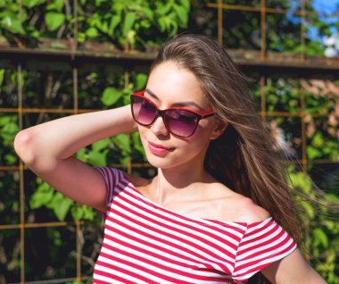Beautiful girl in a bright red T-shirt, fashion lifestyle, posing against the backdrop of fence with green leaves