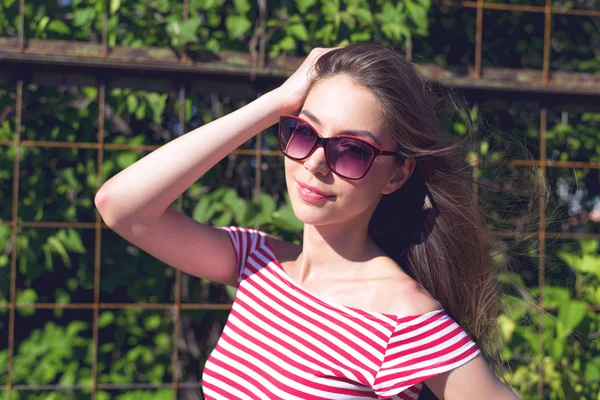 Bella ragazza in una t-shirt rossa brillante, stile di vita di moda, in posa sullo sfondo della recinzione con foglie verdi — Foto Stock