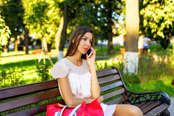 Mooi meisje zittend op een bankje, brunette in roze jurk, fashion lifestyle, praten met de telefoon, luisteren bericht, zonnige dag in het Park, ontspannen, na het werk zakelijke vrouw. — Stockfoto