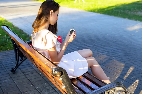 Hermosa chica morena escribiendo un mensaje teléfono parque sentado en el banco en el vestido, mujer de negocios día de verano relajarse después del trabajo, la vida urbana en las redes sociales leer la fuente de noticias . —  Fotos de Stock