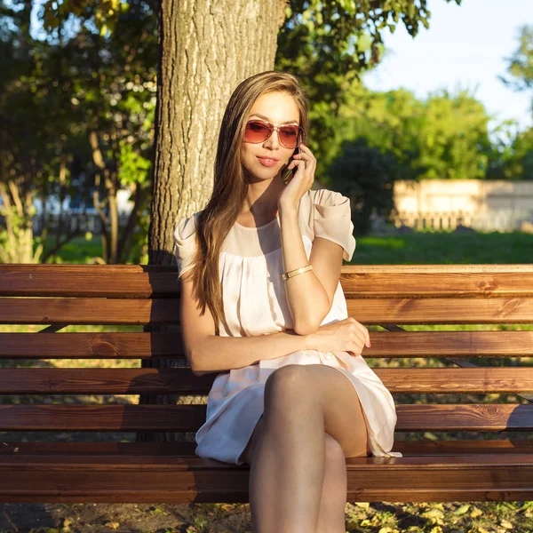 Hermosa chica morena emocional riendo por teléfono en el parque gafas de verano vestido rosa, disfrutar de sus vacaciones en la ciudad. Estilo de vida de moda, un día brillante . — Foto de Stock