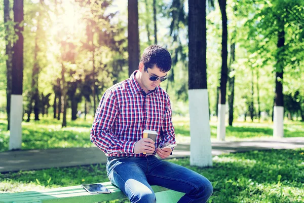 Pria di musim panas di taman, bersantai dengan secangkir kopi atau teh, membaca sms di telepon memakai kacamata, berita feed di jejaring sosial, dalam jins cerah hari. Ide konsep pengusaha — Stok Foto