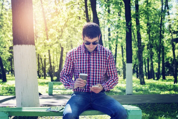 Der Mann im Sommer im Park, entspannt sich bei einer Tasse Kaffee oder Tee, liest SMS am Telefon mit Brille, Nachrichtenfeeds in sozialen Netzwerken, in Jeans strahlend sonnigen Tag. Konzept Geschäftsmann — Stockfoto