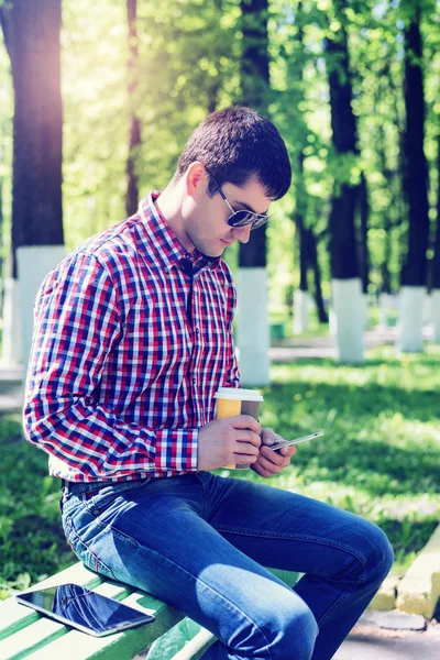 Der Mann im Sommer im Park, entspannt sich bei einer Tasse Kaffee oder Tee, liest SMS am Telefon mit Brille, Nachrichtenfeeds in sozialen Netzwerken, in Jeans strahlend sonnigen Tag. Konzept Geschäftsmann — Stockfoto