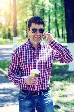 telefonu, akıllı telefon, yaz, işadamı tatil kavramı üzerinde konuşan adam bardaklarda, tişört ve kot pantolon. Şehir bir yaşam tarzı. Park Caddesi. Happy gülümseyen, duygusal. Onun el kupa