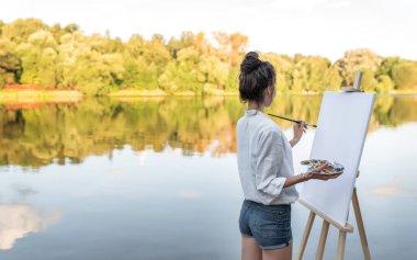 Kız ressam Summer Shore River, bedava uzay mesajı, arka plan su ormanı ağaçları, resim çizmeye başlar, el paletinde boya, fırça sehpası. Beyaz gömlek şortlu. Beyaz boş tuval.