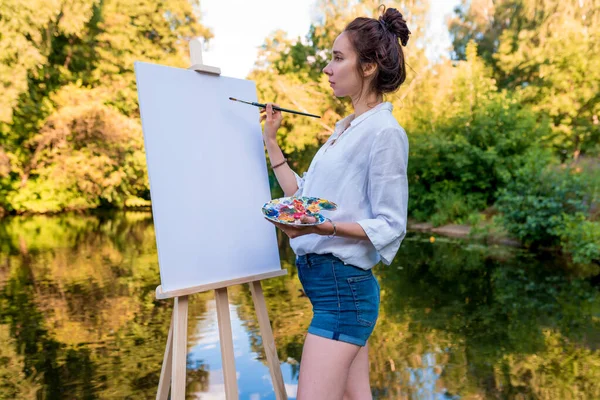 Seniman gadis cantik, taman musim panas di tepi danau, kolam sungai, gambar, palet dengan cat, kuas putih kanvas lukisan, berdiri selondang. Kemeja putih, celana pendek denim. Air latar belakang hutan hijau. — Stok Foto