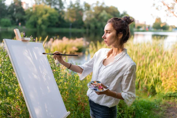beautiful girl, woman artist, in summer on shore of lake, river pond, draws picture, beginning drawing, first brush stroke, brush white canvas painting, easel stand. Green forest trees background.