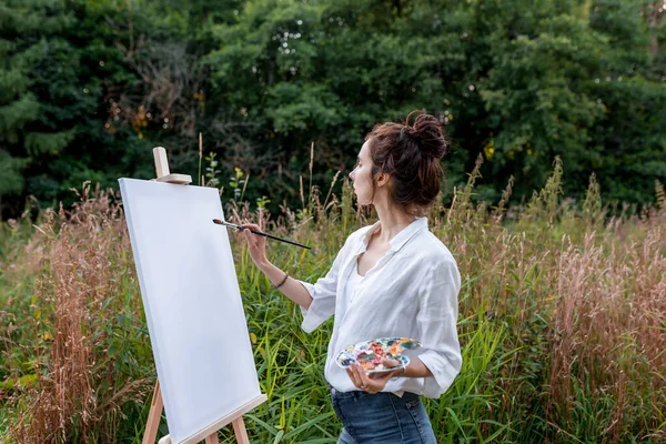 Жінка-художник, влітку в парку кущами, малює картину, палітру з фарбами, пензлем і білим полотном, мольберт. Біла сорочка, джинсові шорти. фон зелених лісових дерев . Стокове Фото