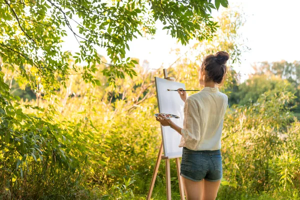 Жінка-художник літній ліс, фонові кущі дерева, починає малювати біле полотно, в кольоровій палітрі ручної щітки. Вільний простір вид ззаду початок малювання, створення креативності та ідеї . Ліцензійні Стокові Зображення