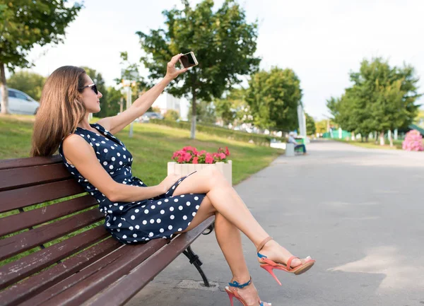 Wanita berpakaian, kota musim panas duduk di bangku mengambil selfie dirinya smartphone. Di telepon genggamnya, rekaman video call. Rambut panjang, pohon latar belakang halaman dan jalan, ruang kosong untuk teks. — Stok Foto