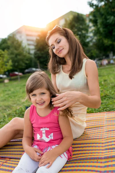 Žena maminka, narovnává vlasy pro dítě, holčička 5-7 let, směje se, usmívá, radostný veselý, venkovní rekreace o víkendu. V létě sedí na dece. Stromy a zelená tráva pozadí. — Stock fotografie