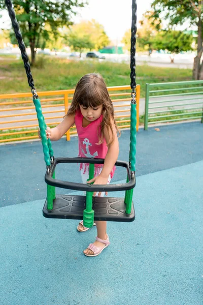Gadis berusia 5-7 tahun, di musim panas di taman bermain, duduk di ayunan anak-anak di taman bermain. — Stok Foto
