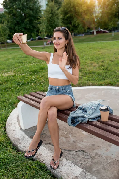 Mujer en verano de la ciudad, se sienta en el banco, agita la mano a sus amigos, grabación de vídeo onlan en el teléfono inteligente. Feliz sonrisa. Tiene un smartphone en la mano. Solicitud de conferencias en línea en Internet. — Foto de Stock