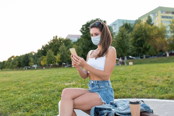 Γυναίκα καλοκαίρι της πόλης, κάθεται πάγκο ιατρική μάσκα, βιντεοκλήση, σε απευθείας σύνδεση εγγραφή βίντεο στο smartphone. Καραντίνα στην πόλη, κλείδωμα. Κρατάει ένα smartphone στο χέρι. Εφαρμογή για online συνέδρια στο Διαδίκτυο. — Φωτογραφία Αρχείου