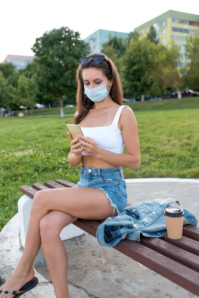 Nyáron egy női város, orvosi maszkban ül egy padon, üzeneteket olvas és ír egy csevegésben. Karantén a városban, lezárás. Egy okostelefont tart a kezében. Online alkalmazás az interneten. — Stock Fotó