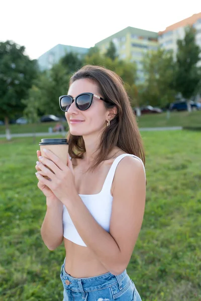 Красива жінка влітку в парку, щаслива усмішка, тримає чашку кави з чаєм в руці, в сонцезахисних окулярах, насолоджуючись запахом і ароматом напою. Фон зелені дерева і трава, будівлі . — стокове фото