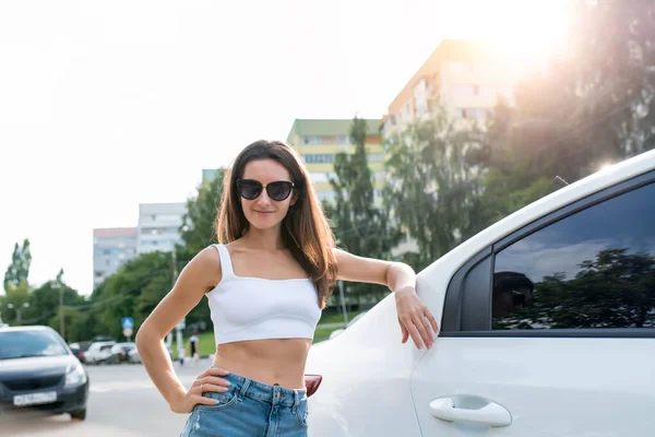 Wanita di musim panas berdiri di samping mobil, sedan putih, seorang gadis cantik tersenyum, kacamata hitam panjang. Denim celana pendek dan atas putih. Stok Foto Bebas Royalti