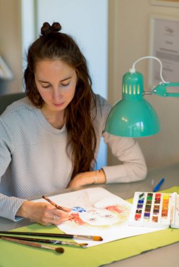 Masadaki kadın, resim çiziyor, kız portresi çiziyor. Işıklı lamba, uzun saçlı. Parlak turuncu boyalar, renk paleti. Siyah masa, fırça boyası. Ev stüdyosu yaratıcılık sanatı kavramı.