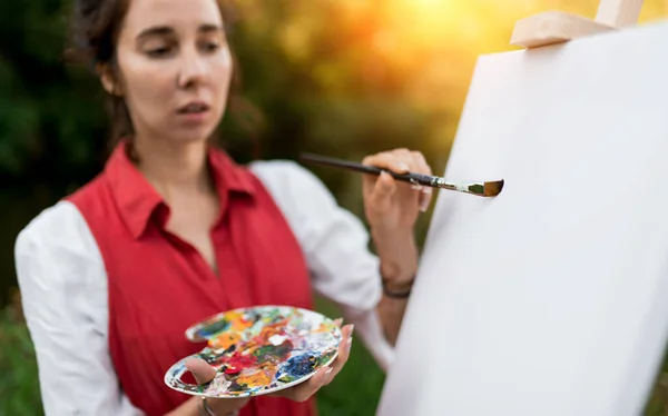 Blurred face out of focus. Close-up of a woman artist, applying first brush stroke to picture with a brush. Colored palette and paintbrush. White picture. The beginning of creativity.