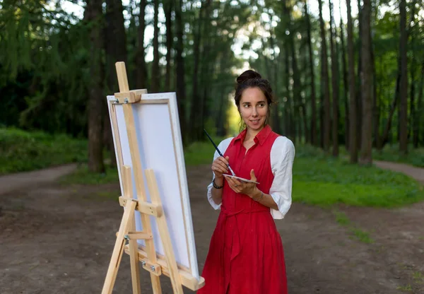 Hermosa artista chica en verano en el bosque, dibuja cuadro, paisaje de fondo y la creación de creatividad. Pintura y caballete de pie. Feliz sonrisa, disfrutando de actividades al aire libre. —  Fotos de Stock