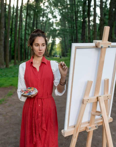 Wanita cantik seniman musim panas di hutan, menggambar gambar, latar belakang pemandangan menciptakan kreativitas. Melukis berdiri selangkangan. Baju merah putih, gaya pohon latar belakang pakaian surut ke kejauhan. Stok Foto