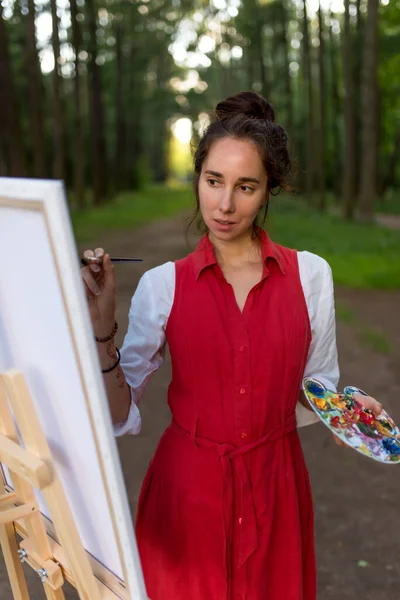 Красива дівчина-художник в літньому лісі, крупним планом, малює картину, ландшафтний фон створення творчості. Живопис мольберт. Стильна червона сукня біла сорочка, кольорова палітра з пензлем . Стокове Фото