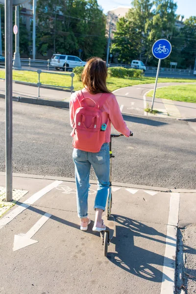 Kvinna i staden på sommaren står vid ett vägskäl med en skoter. Vi väntar på att trafiksignalen ska korsa vägen. Jeans jacka och ryggsäck bakom ryggen. — Stockfoto