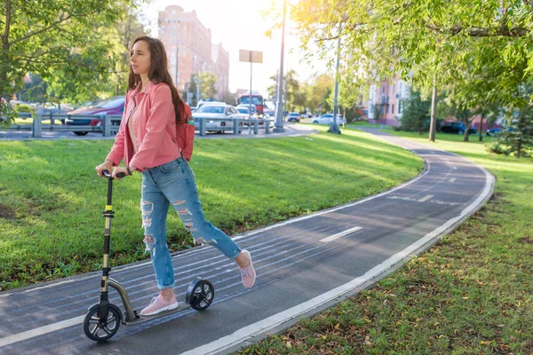 Menina no verão na cidade monta uma scooter, em jeans de roupa casual e uma jaqueta rosa, espaço livre de uma cópia do texto. Fundo estrada verde grama e árvores, rodovia e carros. — Fotografia de Stock