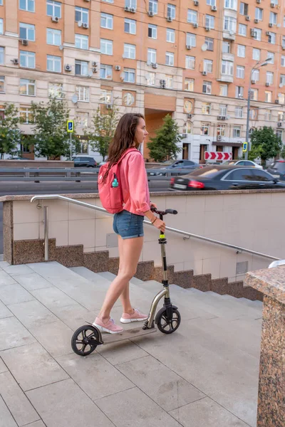Woman in summer with a scooter, descends into underground pedestrian to cross road, background road cars, buildings and steps into crossing. Denim shorts pink jacket and backpack. — Stock Photo, Image