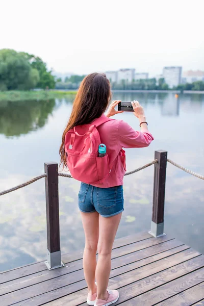 Жінка влітку в місті поблизу озера, річкового ставка, фотографує ландшафтний смартфон, записує відео Інтернет, прикладну камеру смартфона. Водні дерева фон пірс дріб'язковий. Повсякденний одяг рожева куртка . — стокове фото