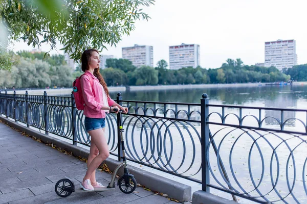 Woman in summer in city near lake, river pond, looks at landscape, scooter, pink backpack behind her back, free space for copy text. Relaxation after work, enjoying nature. — Stock Photo, Image