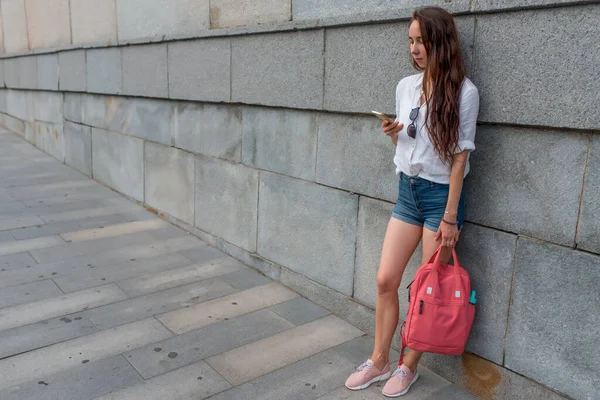 Дівчина в місті в літню дату, читає повідомлення і пише текст в додатку, онлайн в смартфоні. Сумка рюкзак, біла сорочка та рожевий рюкзак. Фонова стіна переходить дорогою. Вільний простір для тексту . — стокове фото
